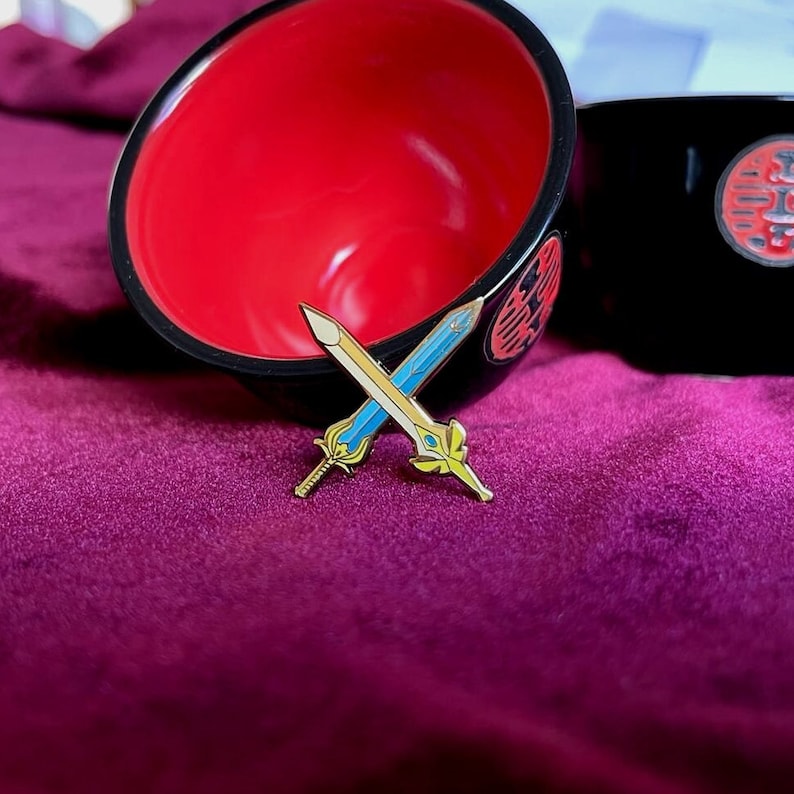 May include: Two crossed sword pins with gold and blue enamel detailing, set against a deep purple background. The pins are in front of a black and red bowl with a decorative design.