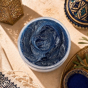 Puede incluir: Un tarro transparente lleno de un producto cosmético azul oscuro, que muestra una textura arremolinada. La imagen incluye elementos decorativos como una linterna dorada y un cuenco de cerámica estampada. El fondo es una superficie de color beige claro.