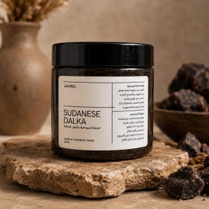 Peut inclure: Un pot cylindrique avec un couvercle noir contenant une pâte brune foncée Sudanese Dalka. L'étiquette blanche porte le texte LAUREL, SUDANESE DALKA, MADE BY SUDANESE HANDS et 200g, ainsi que du texte arabe détaillant les bienfaits et l'utilisation.