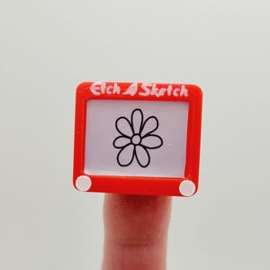 May include: A red plastic toy Etch A Sketch with a white flower drawn on the screen. The toy has white knobs on the sides.