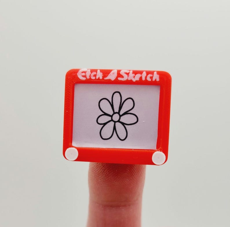May include: A red plastic toy Etch A Sketch with a white flower drawn on the screen. The toy has white knobs on the sides.