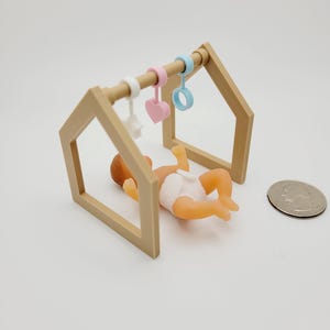 May include: A wooden baby play gym with a hanging heart, star, and ring. A small doll is lying on its back inside the gym.