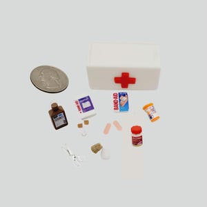 May include: A miniature white first aid kit with a red cross, surrounded by small medical supplies. Includes a bottle of medicine, bandages, pills, scissors, and a quarter for scale. The Band-Aid box is visible.