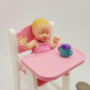 May include: A miniature doll of a baby in a pink high chair with a tray. The baby doll is wearing a pink dress and has blonde hair. A small teal sippy cup with a purple lid sits on the tray. The high chair is pink and white.