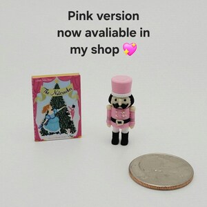 May include: A miniature book titled The Nutcracker with a pink and blue cover featuring a Christmas tree, alongside a small pink nutcracker figurine and a US quarter coin. Text reads: Pink version now avaliable in my shop.