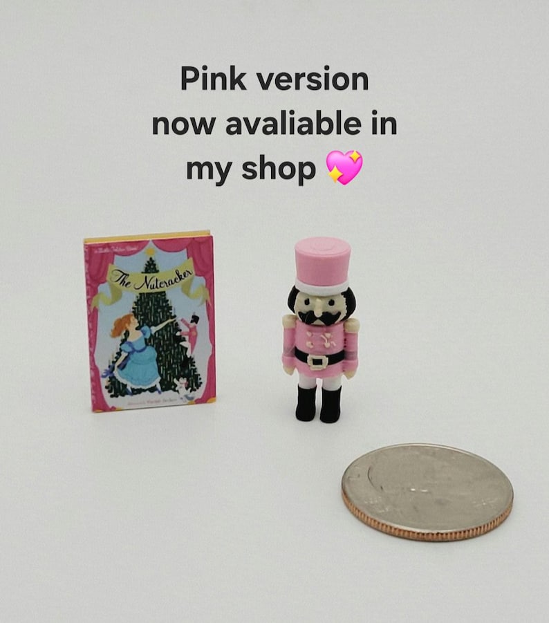 May include: A miniature book titled The Nutcracker with a pink and blue cover featuring a Christmas tree, alongside a small pink nutcracker figurine and a US quarter coin. Text reads: Pink version now avaliable in my shop.