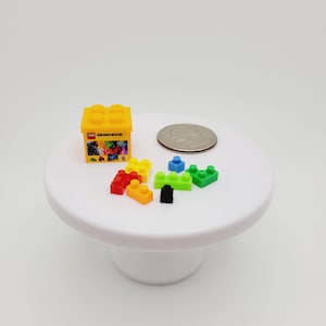 May include: A small yellow box with the words "Mini Building Blocks" printed on the side. The box is open and contains a variety of colorful plastic building blocks. There is a quarter next to the box for scale.