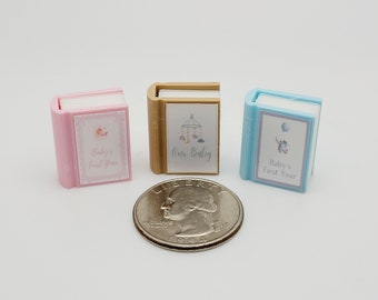 May include: Three miniature baby books with different colored covers. The pink book says "Baby's First Year", the gold book says "Our Baby", and the blue book says "Baby's First Year".