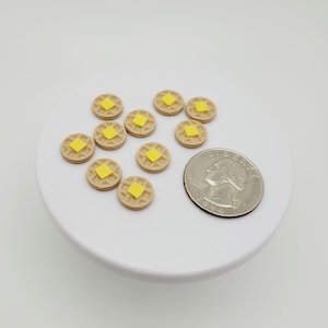 May include: Miniature waffles with yellow butter squares on a white surface. The waffles are light brown with a waffle pattern. A quarter is next to the waffles for size comparison. The waffles are likely for dollhouses or crafts.