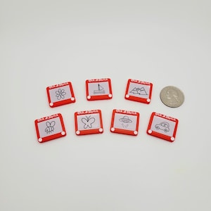 May include: Eight red Etch A Sketch toys with different drawings on the screen. The drawings include a flower, a sailboat, mountains, a bee, a butterfly, a UFO, and a car.