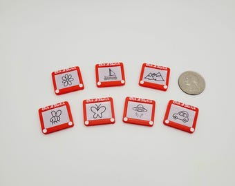 May include: Eight red Etch A Sketch toys with different drawings on the screen. The drawings include a flower, a sailboat, mountains, a bee, a butterfly, a UFO, and a car.