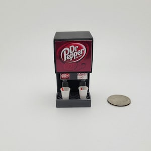 May include: Miniature Dr Pepper soda fountain with the Dr Pepper logo on the front. The fountain has two dispensers and two small paper cups. A coin is next to the fountain.