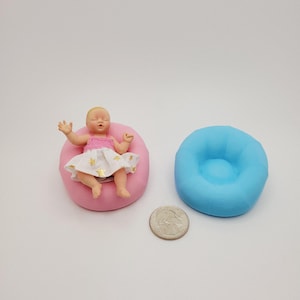 May include: A miniature doll wearing a pink top and a white skirt with gold stars, resting on a pink beanbag chair. A blue beanbag chair and a quarter are also present, suggesting a dollhouse or miniature theme.