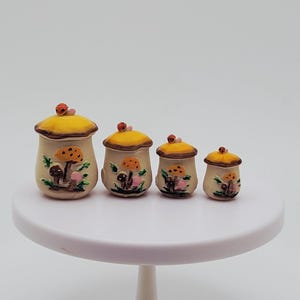 May include: A set of four graduated beige canisters with yellow mushroom-shaped lids. Each canister features a painted orange mushroom design on the front and a small ladybug figurine perched on top of the lid, arranged from largest to smallest.