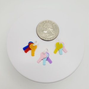 May include: A collection of colorful miniature key charms in red, orange, blue, yellow, pink, light blue, and purple. A US quarter is next to the charms for size comparison. The keys are on a white circular surface.