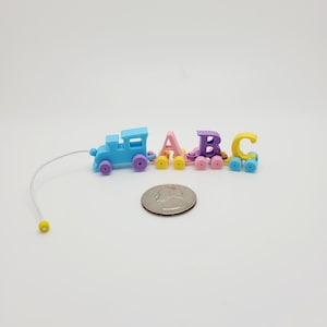 May include: A miniature toy train set with the letters A, B, and C. The train is light blue with purple wheels, and the letters are pastel colors. A quarter is next to the train for size comparison.