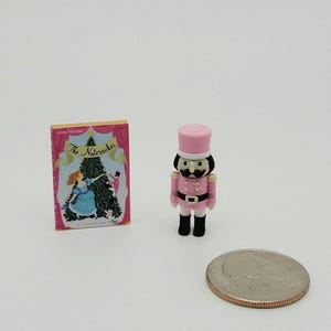 May include: A miniature book titled The Nutcracker with a colorful cover illustration of a Christmas tree and a small pink nutcracker figurine standing next to a United States quarter coin for scale, showing the relative size of these tiny items.