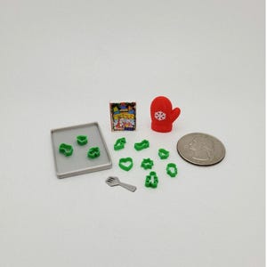 May include: Miniature baking set with green cookie cutters in various shapes, a red mitten with a snowflake, a quarter, a small cookbook, a gray baking sheet, and a spatula. The set is designed for dollhouses or craft projects.