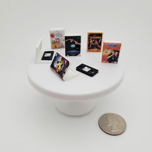 May include: Miniature VHS tapes and movie cases, including "Top Gun" and "The Goonies," are arranged on a white cake stand. A quarter is placed nearby for scale. The movie cases are colorful, and the VHS tapes are black.