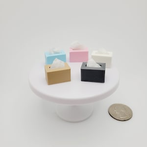 May include: Six miniature tissue box holders in different colors: light blue, pink, white, brown, black, and gray. The boxes are arranged on a white cake stand with a silver coin in the background.