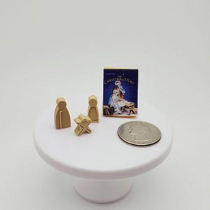 May include: Miniature Christmas scene on a white pedestal. Includes wooden figures, a star, and a small book titled "The Christmas Story." A quarter is included for scale. The book cover features an illustration of the nativity scene.