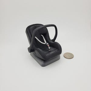 May include: A black miniature car seat with a white strap and buckle. The seat has a handle and is sitting on a gray base. A quarter is next to the seat for size comparison.