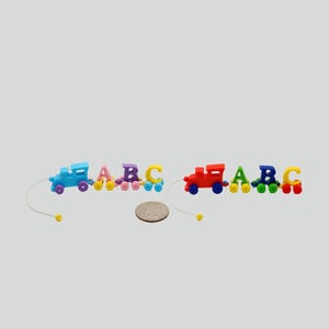 May include: Two miniature wooden train sets, each featuring a locomotive followed by the letters A, B, and C on wheels. The left train is blue, pink, purple, and yellow. The right train is red, green, dark blue, and yellow. A US quarter coin is placed between them.