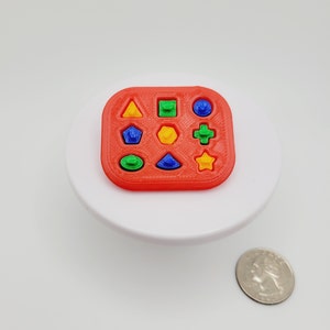 May include: A red shape sorter toy with nine different colored shapes: a yellow triangle, green square, blue circle, green oval, yellow hexagon, blue triangle, green cross, and yellow star. A quarter is next to the toy.