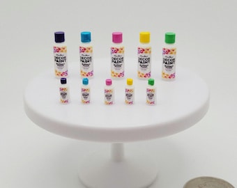 May include: A collection of miniature paint bottles with colorful caps, including purple, blue, pink, yellow, and green. The bottles are labeled "Decor Paint" and are arranged on a white cake stand. A quarter coin is next to the stand.