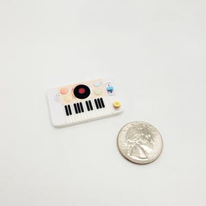 May include: A miniature white toy keyboard with black keys, colorful buttons, and a record design. A US quarter is next to the toy for size comparison. The toy is on a white background.