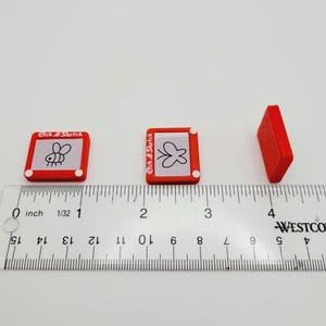 May include: Three red Etch A Sketch toys, each with a different drawing on the screen. The toys are lined up on a white surface with a ruler underneath. The ruler is marked in inches.