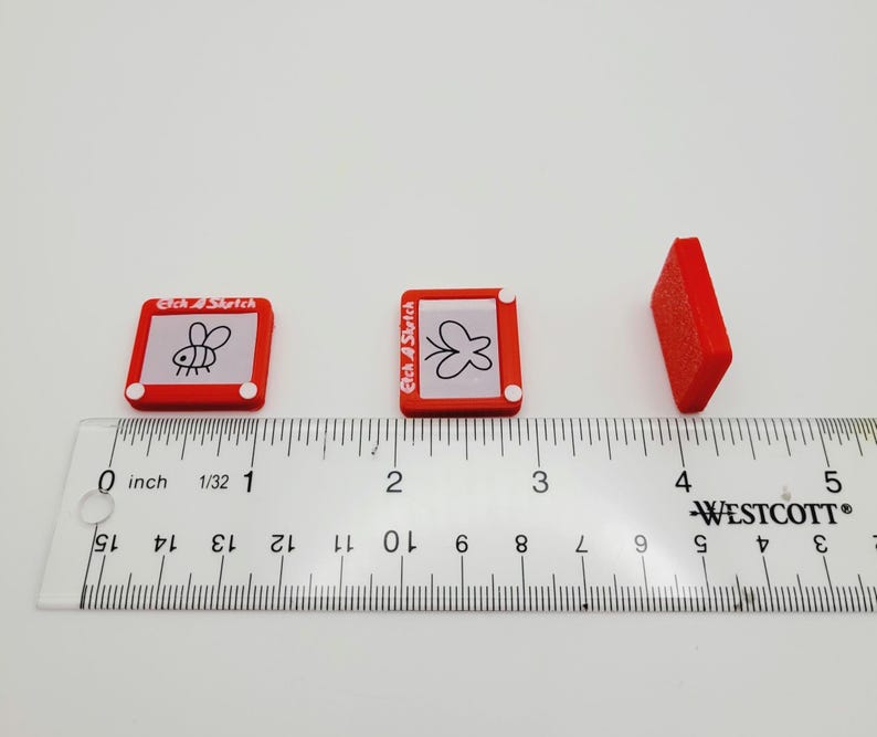 May include: Three red Etch A Sketch toys, each with a different drawing on the screen. The toys are lined up on a white surface with a ruler underneath. The ruler is marked in inches.