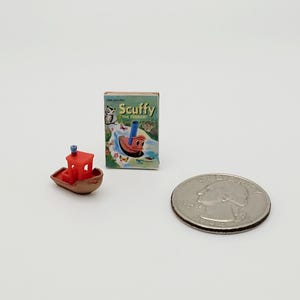 May include: A miniature red tugboat with a blue smokestack, a brown hull, and a small white window. The tugboat is next to a small book titled "Scuffy the Tugboat" and a US quarter for scale.