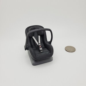 May include: A small, black infant car seat with white straps and a buckle. The seat has a handle and is set on a rectangular base. A coin is next to the seat for size comparison.