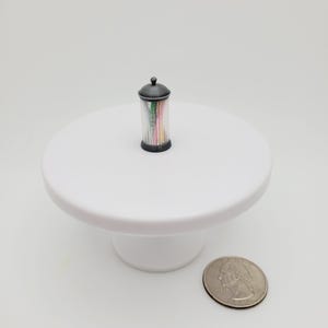 May include: A small black and clear glass jar with a lid filled with colorful toothpicks. The jar is sitting on a white cake stand.