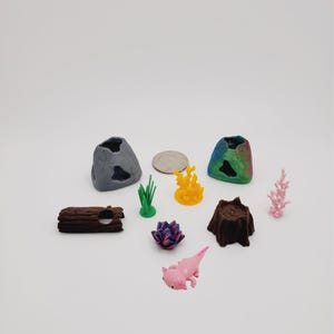 May include: A set of miniature aquarium decorations, including a gray and a rainbow-colored cave, a brown log, a green plant, a yellow coral, a pink coral, a pink flower, a pink axolotl, and a brown tree stump.