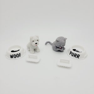 May include: White plastic dog and cat food bowls with black paw print and fish bone designs. The bowls say "WOOF" and "PURR". A white dog and gray cat figurine are next to the bowls.