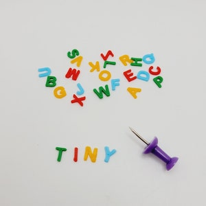 May include: A white surface with colorful plastic letters spelling out the word "TINY" and a purple push pin. The letters are scattered around the word "TINY" and include the letters A, B, C, D, E, F, G, J, K, L, M, N, O, P, R, S, T, U, W, X, Y, and Z.