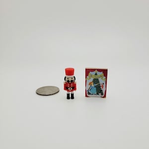 May include: Miniature toy soldier in a red uniform and black boots, with a red hat. Beside it is a tiny book titled "The Nutcracker" with a festive illustration. A coin is also present.