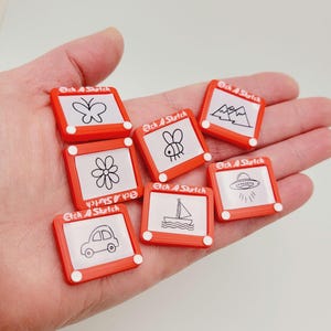 May include: Eight red Etch A Sketch toys with different drawings on the screen. The drawings include a butterfly, mountains, a bee, a flower, a sailboat, a UFO, and a car.