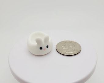 May include: A small white ceramic bunny-shaped dish with black eyes and pink cheeks. The dish is about the size of a US quarter.