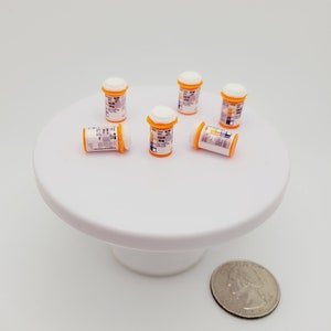 May include: Six miniature plastic pill bottles with white lids and labels. The bottles are arranged on a white cake stand. A US quarter is in the foreground for scale.