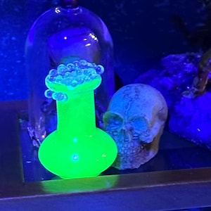 May include: A green glass bottle with a round bottom and a narrow neck, filled with small white beads, sits under a glass dome. A white skull sits next to the bottle on a dark surface.