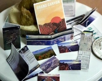 May include: Miniature scene of a white table with a collection of tiny travel souvenirs. A "Grand Canyon" booklet is visible, along with other small brochures and a US quarter. The scene is set on a blue patterned rug, creating a travel-themed display.