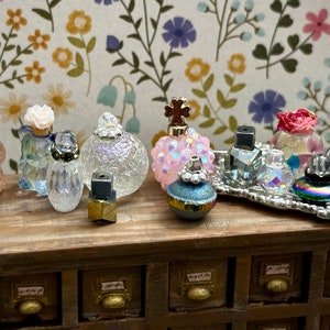 May include: A collection of miniature perfume bottles with various designs and colors, including clear glass, iridescent, and pink. Some bottles have flower accents and others have crystal-like stoppers. The bottles are arranged on a wooden surface with a silver tray holding several bottles.