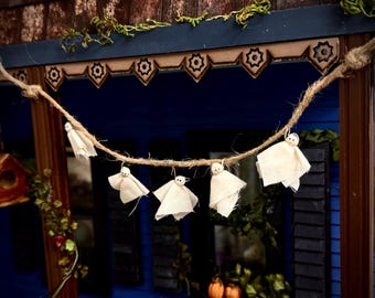 May include: A decorative garland featuring five small ghost figures made of off-white fabric draped over a thick, natural-colored jute twine. Each ghost has a small, round, light-colored head with two tiny black dots for eyes, suspended from the rope.