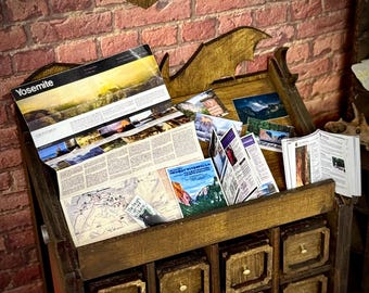 May include: A miniature wooden display cabinet filled with brochures, maps, and postcards. The cabinet has multiple drawers and a decorative top. The brochures feature images of Yosemite National Park. The cabinet is set against a red brick wall.