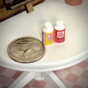 May include: Two miniature bottles of Mod Podge adhesive, one with a yellow label and one with a red label, positioned next to a silver-coloured Cumberland Island 2018 quarter coin for scale, showing the small size of the craft supplies.