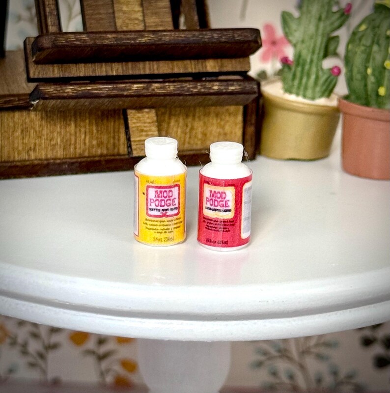May include: Two miniature bottles of Mod Podge adhesive. One bottle features a yellow label with black and pink text, while the other bottle features a red label with black and white text. Both bottles have white screw-on caps and cylindrical bodies.