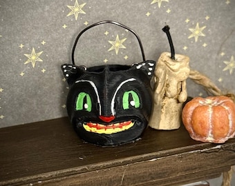May include: A miniature black cat bucket with a metal handle, featuring bright green eyes, a red nose, and a yellow and red mouth. Beside the bucket sits a small, off-white textured candle with a black wick and a tiny orange pumpkin figurine.